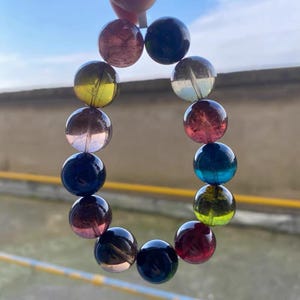 Puede incluir: Una pulsera compuesta por una serie de cuentas esféricas, coloridas y translúcidas. Las cuentas son en tonos de rojo, azul, amarillo y transparente, ensartadas para formar una forma circular. La pulsera se sostiene sobre un fondo de cielo azul.