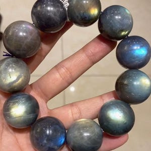 May include: A bracelet composed of eleven polished, spherical labradorite beads. Each bead displays a dark gray base color with iridescent flashes of blue, gold, and green light reflecting from the surface. A small white tag with black text is attached.