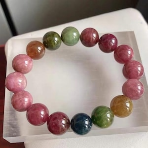 Puede incluir: Un brazalete de cuentas con una variedad de piedras preciosas redondas y coloridas. Las cuentas son en tonos de rosa, verde, marrón y azul oscuro. El brazalete se exhibe sobre un bloque rectangular transparente.