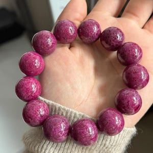 May include: A bracelet with large, round beads in a deep magenta colour. The beads have a polished, glossy finish, and the bracelet is displayed on a person's wrist, showcasing the jewellery's colour and texture. The beads appear to be approximately 2.5 cm in diameter.
