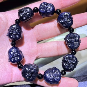 Puede incluir: Un brazalete azul oscuro con caras talladas del Buda de la Risa. El brazalete está hecho de cuentas de piedra pulidas con acentos negros.