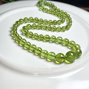 Puede incluir: Collar de cuentas redondas y translúcidas, de color verde claro. Las cuentas están ensartadas, formando una curva sobre un plato blanco. Las cuentas varían en tamaño, siendo las más grandes las del centro.