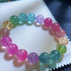 Puede incluir: Una pulsera de cuentas coloridas con cuentas redondas translúcidas en tonos rosa, morado, verde, azul y amarillo. La pulsera se exhibe sobre una superficie blanca, mostrando los colores vibrantes de las piedras preciosas.