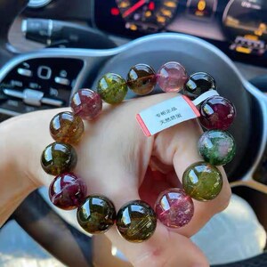 Puede incluir: Un brazalete hecho de piedras preciosas redondas multicolores. El brazalete está hecho de piedras preciosas verdes, rosas y negras. El brazalete está en la muñeca de una persona. La etiqueta en el brazalete dice "天然碧玺".