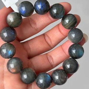 May include: A beaded bracelet composed of round, polished labradorite stones. Each dark grey bead displays iridescent flashes of blue and gold light, reflecting a smooth, reflective surface texture throughout the circular arrangement of the jewelry.