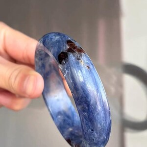 以下が含まれることがあります： 滑らかで丸みを帯びた表面を持つ、磨かれたブルーカイヤナイトのバングルブレスレット。半透明の青い石の構造の中に、繊維状の針のようなインクルージョンと、濃い赤褐色の鉱物の斑点がはっきりと見られます。
