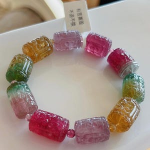 Puede incluir: Un brazalete hecho de cuentas de piedras preciosas talladas multicolores. Las cuentas son cilíndricas y tienen un patrón tallado en ellas. El brazalete está sobre una superficie blanca con una etiqueta que dice "特级翡翠 水润不燥".