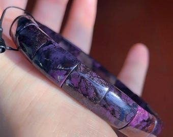 Puede incluir: Una pulsera elástica compuesta por cuentas rectangulares pulidas de charoíta morada y negra. Las cuentas presentan un patrón natural veteado con inclusiones de color violeta intenso y gris oscuro, ensartadas en un fino cordón elástico negro.