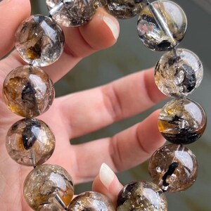 Peut inclure: Un bracelet composé de perles rondes et polies en quartz clair, présentant des inclusions visibles de dépôts minéraux brun foncé, noirs et jaune doré. Les perles sont enfilées sur un cordon flexible, formant une structure circulaire lisse.
