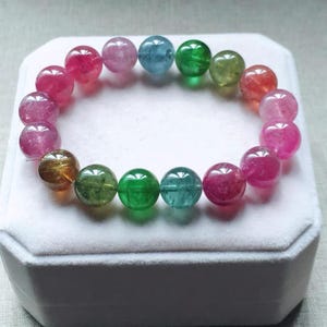 Puede incluir: Un colorido brazalete hecho de piedras preciosas redondas y pulidas. Las cuentas son una mezcla de tonos rosas, verdes, azules y amarillos. El brazalete se exhibe sobre una superficie blanca.