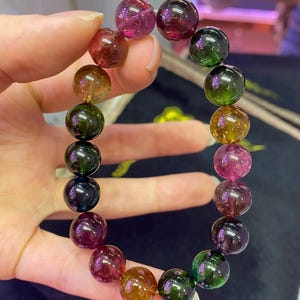 Puede incluir: Un brazalete de piedras preciosas multicolores con una variedad de cuentas redondas. Las cuentas muestran una gama de colores, incluyendo rosa, verde, amarillo y marrón. El brazalete se sostiene en una mano, mostrando las piedras pulidas y translúcidas.