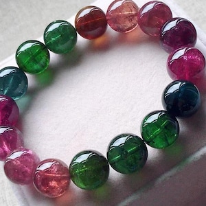 Puede incluir: Un brazalete compuesto por piedras preciosas redondas y pulidas en tonos verdes, rojos y morados. Las cuentas están ensartadas, formando una forma circular. Las piedras preciosas tienen una superficie brillante y reflectante.