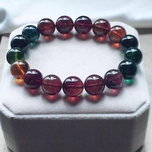 Puede incluir: Un brazalete de cuentas con cuentas de piedras preciosas en tonos rojos, verdes y naranjas. Las cuentas redondas están ensartadas, creando un diseño circular. El brazalete se exhibe sobre una superficie blanca.