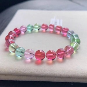 Puede incluir: Un brazalete colorido con una variedad de piedras preciosas redondas y pulidas. Las cuentas muestran un espectro de colores, incluyendo rojo, rosa, verde y transparente, creando un accesorio vibrante y llamativo. El brazalete se exhibe sobre una superficie neutra.