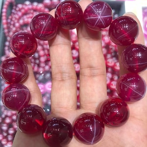 Puede incluir: Un brazalete de piedras preciosas rojas con inclusiones en forma de estrella. El brazalete está hecho de cuentas redondas y está enhebrado en un cordón elástico transparente.