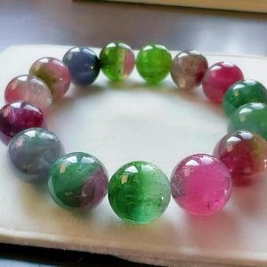 Puede incluir: Un brazalete con grandes cuentas de piedras preciosas pulidas dispuestas en círculo. Las cuentas muestran una variedad de colores, incluyendo verde, rosa, morado y azul. El brazalete está sobre un fondo blanco.