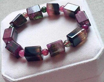 Könnte beinhalten: Ein Armband aus facettierten, quadratischen, mehrfarbigen Edelsteinen. Das Armband zeigt eine Mischung aus schwarzen, grünen, rosa und violetten Farbtönen. Die Edelsteine sind mit kleinen, silberfarbenen Perlen aufgereiht.