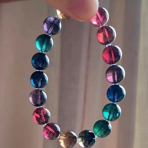 Puede incluir: Un brazalete de cuentas con una variedad de piedras preciosas redondas y coloridas. Las cuentas son de color rojo, rosa, verde, azul, morado y dorado. El brazalete está sostenido por un dedo.