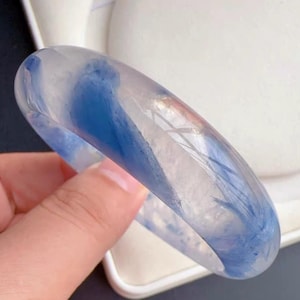 May include: A translucent, rounded bangle bracelet featuring a smooth, polished surface with swirling inclusions of deep blue and white patterns throughout the clear material, creating a distinct visual depth within the circular band structure.