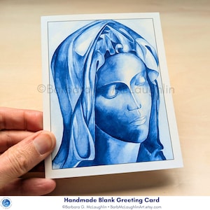 A handmade greeting card with an envelope. The card features a print of my blue ink painting the Blessed Virgin Mary inspired by Michelangelo's Pieta. The Virgin Mary's face is serene, with closed eyes, and detailed veil. The handmade blank greeting card is held in my hand.