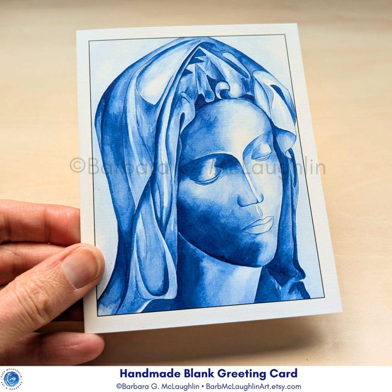 A handmade greeting card with an envelope. The card features a print of my blue ink painting the Blessed Virgin Mary inspired by Michelangelo's Pieta. The Virgin Mary's face is serene, with closed eyes, and detailed veil. The handmade blank greeting card is held in my hand.