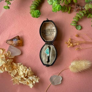 May include: A vintage jewellery box, open to reveal a silver ring with a turquoise triangular stone. The box is black with a cream interior. The background is a pink surface, decorated with dried flowers and greenery.