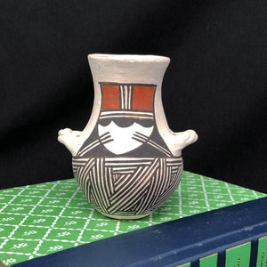 May include: A small, white ceramic vase with black and red geometric designs. The vase has two small handles on the sides and is sitting on top of a green book.