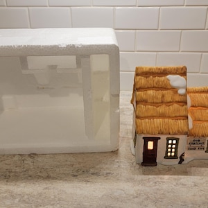 Puede incluir: Una casa de cerámica de un pueblo navideño con tejado de paja, detalles de nieve y ventanas iluminadas. La casa tiene el texto "Old Country Inn" y está junto a una caja de poliestireno blanca.