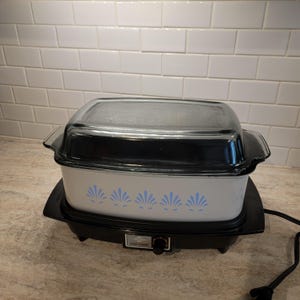 Puede incluir: Una olla de cocción lenta rectangular blanca con base negra y tapa de cristal transparente. El exterior blanco presenta un patrón floral azul en forma de abanico. Un mando de control negro se sitúa en la parte frontal con el texto SLOW COOKER.