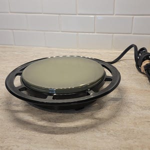 May include: A round electric mug warmer featuring a smooth, light gray heating surface surrounded by a black circular frame. The device includes a black power cord with a standard two-prong plug, coiled and secured with a small piece of tan tape.
