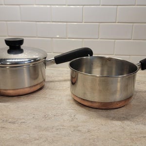 May include: Two stainless steel saucepans with copper-coloured bases and black handles. One saucepan has a lid with a black knob. The pans are on a light-coloured countertop, suitable for cooking.