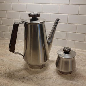 May include: A stainless steel coffee pot and sugar bowl set. The coffee pot has a long spout and a dark brown handle. The sugar bowl has a matching lid. Both have dark brown accents on the lids.