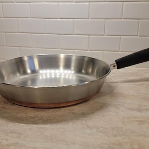 May include: A stainless steel frying pan with a black handle. The pan has a copper-colored base and is sitting on a light-colored countertop. The background features white tiles.