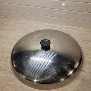 Puede incluir: Una tapa de olla de acero inoxidable con un asa negra. La tapa tiene un diseño de ladrillo y es circular. La tapa está sobre una encimera de color claro.