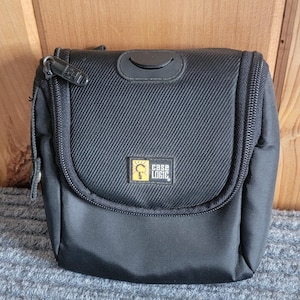 May include: A black, rectangular, zippered carrying case with a textured fabric exterior. The front features a small, square yellow and black logo patch with the text Case Logic. The top flap includes a black, semi-circular rubberized accent.