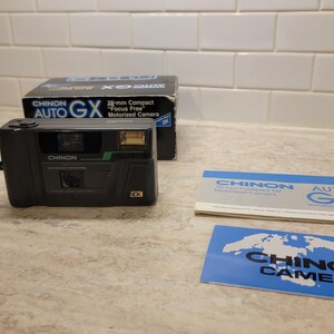 Puede incluir: Una cámara compacta Chinon Auto GX de 35 mm negra y verde con enfoque automático y motorizada, con una correa negra. La cámara está sobre una superficie de azulejos blancos con una caja y manuales de instrucciones.