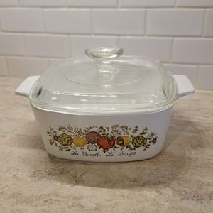 May include: White square casserole dish with a clear glass lid. The dish features a colorful vegetable and herb design with the words "La Parsil, La Sauge" printed on the front. The dish has two white handles.