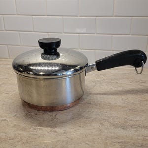 以下が含まれることがあります： 銅底と黒いハンドルが付いたステンレス鋼のソースパン。鍋には黒いノブが付いた蓋が付いています。ハンドルは端にリングが付いています。鍋は明るい茶色のカウンタートップにあります。