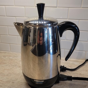 Puede incluir: Una cafetera eléctrica Farberware de acero inoxidable con asa y pomo de tapa negros. La cafetera tiene un pico y una tapa transparente. El cable de alimentación está enchufado.