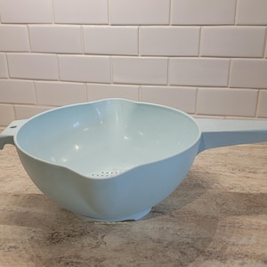 May include: A light blue plastic colander with a handle and a pouring spout. The colander has a rounded bowl shape with small drainage holes at the bottom. It is designed for rinsing and draining food items.