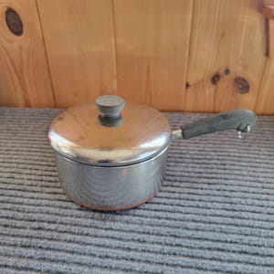 May include: A stainless steel saucepan with a matching lid and a gray handle. The pot features a polished metallic finish, a circular lid with a central gray knob, and a long, ergonomic gray handle with a small metal loop at the end.
