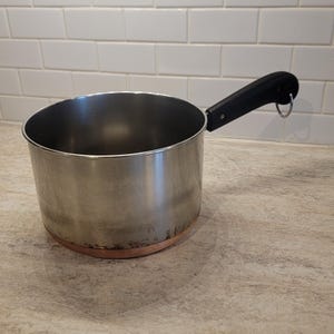 May include: A stainless steel saucepan with a black handle and a copper-coloured base. The pan is on a light-coloured countertop in front of a white tiled wall. The handle has a metal ring.