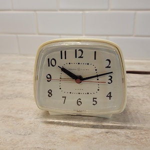 Puede incluir: Reloj despertador General Electric de color crema con una esfera rectangular y esquinas redondeadas. La esfera del reloj tiene números negros, manecillas negras y una manecilla de alarma roja. El texto "GENERAL ELECTRIC" y "ALARM" son visibles.