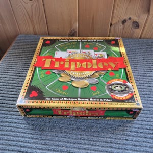 May include: A square Tripoley Special Edition board game box featuring a green playing surface with red heart symbols, playing cards, and gold tokens. The box displays the title Tripoley in gold letters on a red banner and includes a rotating turntable.