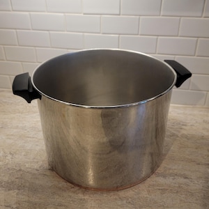 May include: A large, stainless steel cooking pot with black handles. The pot is cylindrical with a wide opening and sits on a countertop. The background features white subway tile.