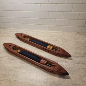 Peut inclure: Deux navettes de tissage en bois brun foncé, lisses et allongées, chacune dotée d'un centre creux contenant une broche métallique entourée de fil bleu foncé. Les navettes ont des extrémités pointues et de petits composants noirs.