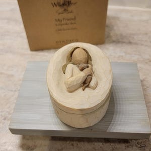 Könnte beinhalten: Eine beige Willow Tree Andenkenbox mit einer geschnitzten Figur einer Person, die ein Tier hält. Die Box steht auf einem grauen Holztablett, mit einem Karton im Hintergrund. Der Text "My Friend" ist sichtbar.