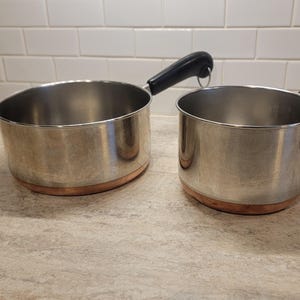 May include: Two stainless steel saucepans with copper bases and black handles. The pans are on a light-coloured countertop. The handles have a loop for hanging.