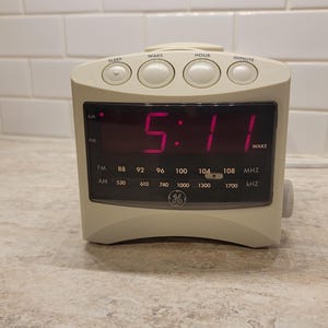 May include: A beige GE digital alarm clock displaying 5:11 in red digits. The clock has buttons for sleep, wake, hour, and minute. It also features FM and AM radio frequency indicators.
