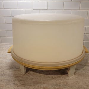 Könnte beinhalten: Ein runder, cremefarbener Kuchenbehälter mit gelbem Sockel und einem transparenten, abgerundeten Deckel. Der Behälter steht auf einem weißen Ständer mit vier Beinen. Der Hintergrund zeigt eine weiße Fliesenspiegel.